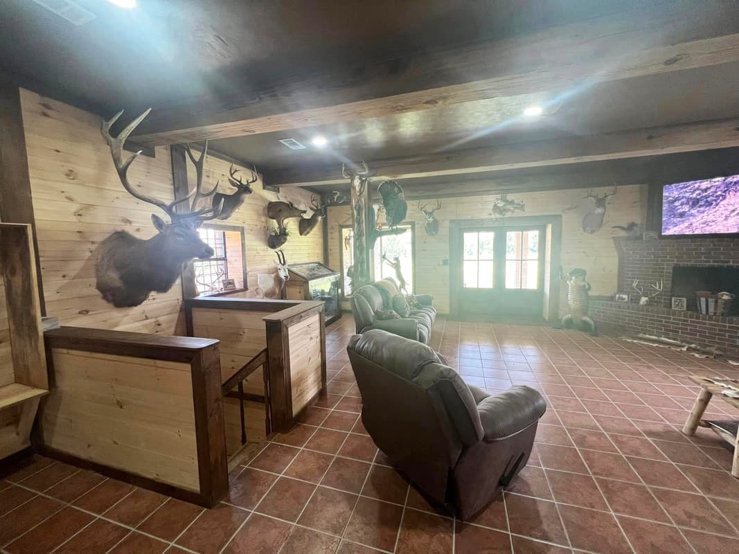 Interior of a rustic hunting lodge living room with wood panel walls, mounted deer trophies on display, leather recliners, and a brick fireplace