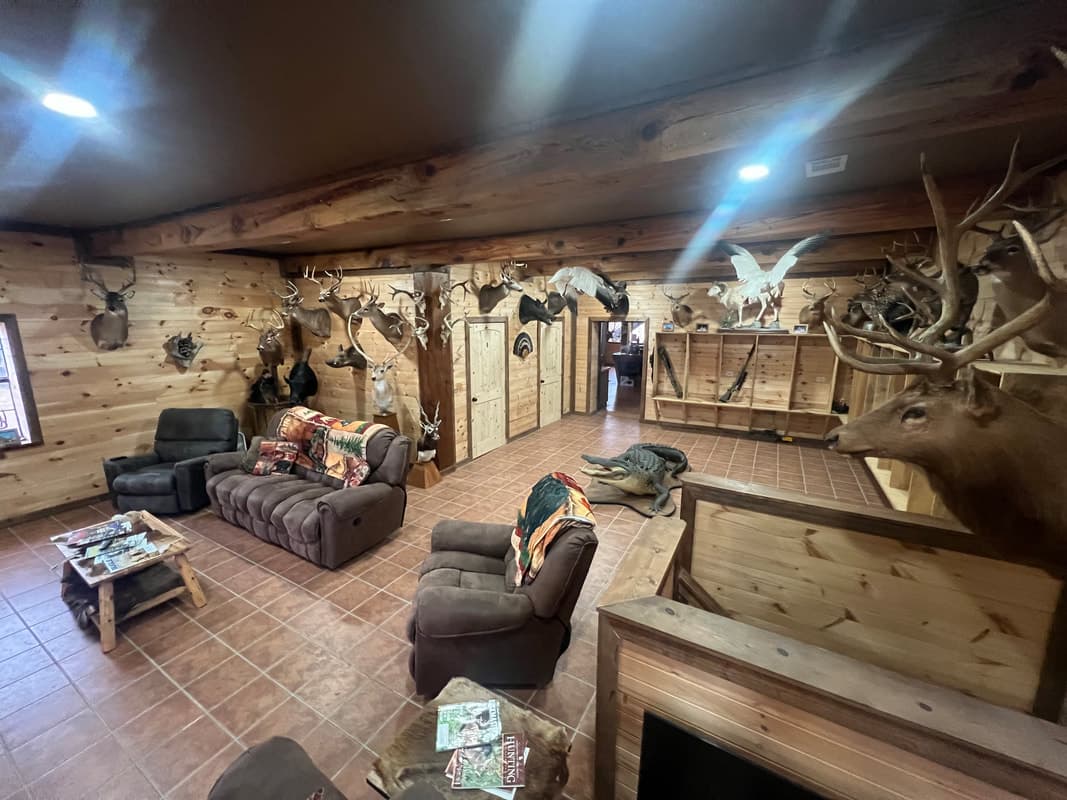 Rustic lodge living room interior with wood paneling, comfy seating, and numerous mounted deer heads along the walls